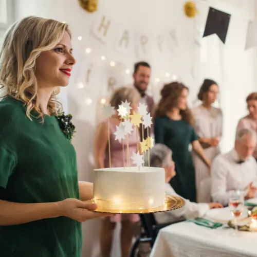 Woman holding a decorated birthday cake at a festive event with guests celebrating, highlighting Homemade By Colleen's catering for family gatherings in Pittsburgh.