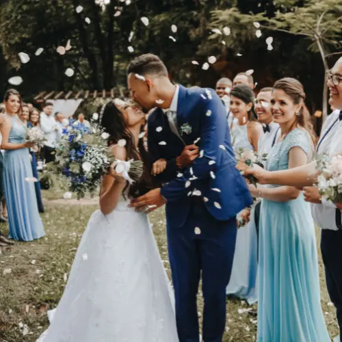 Bride and groom kissing surrounded by wedding guests in blue attire, celebrating with flower petals in a lush outdoor setting, highlighting personalized catering for special events in Pittsburgh.